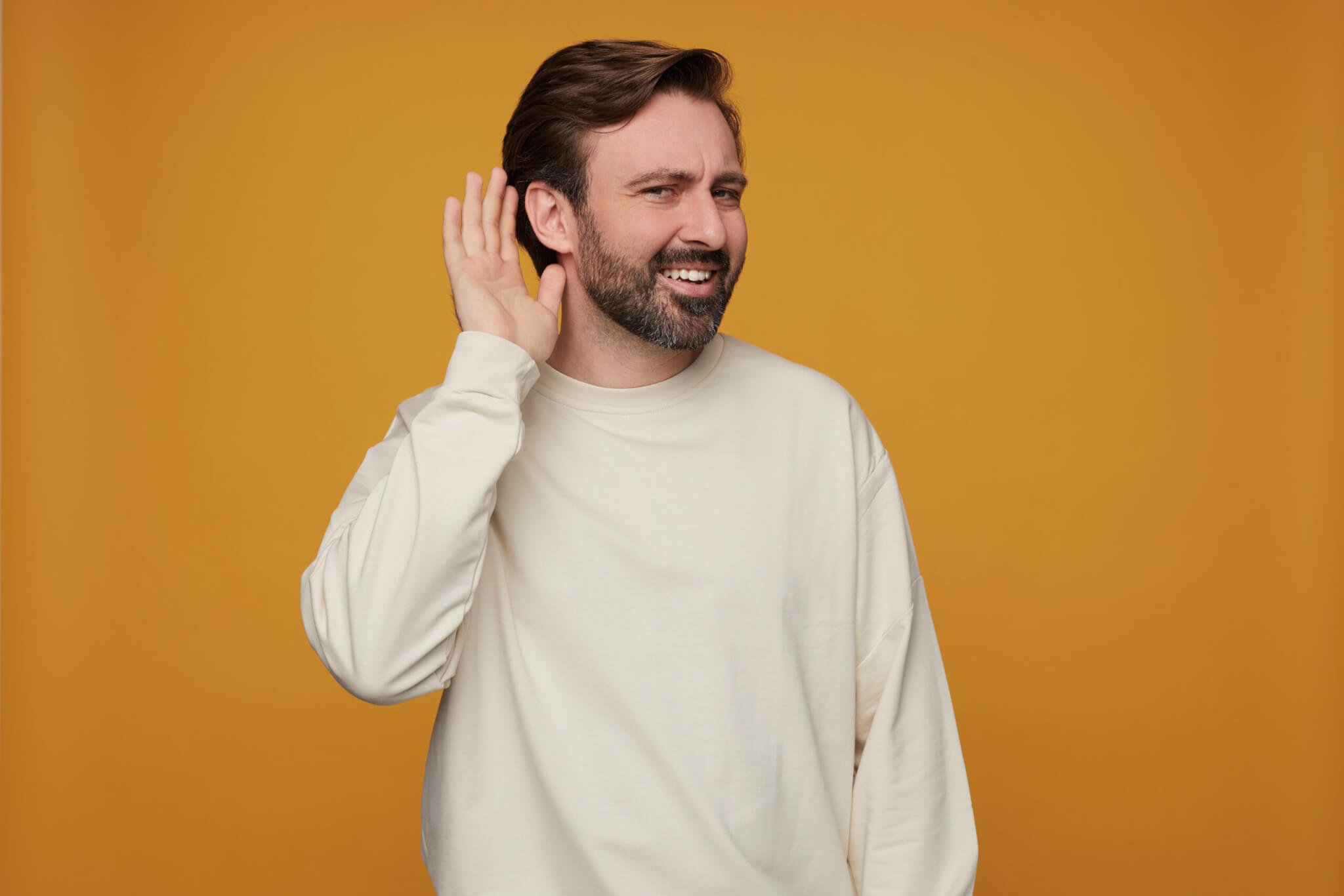 indoor portrait man wears white sweatshirt posing yellow background keeps his hand near ear trying hear something scaled